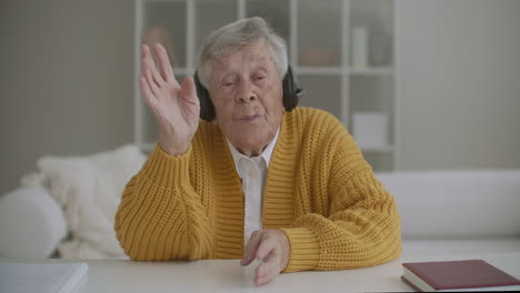 Eine-ältere-Großmutter-Von-80-Jahren-Winkt-Mit-Der-Hand-In-Die-Kamera-Und-Grüßt-Per-Videokommunikation,-Während-Sie-Mit-Kopfhörern-Sitzt